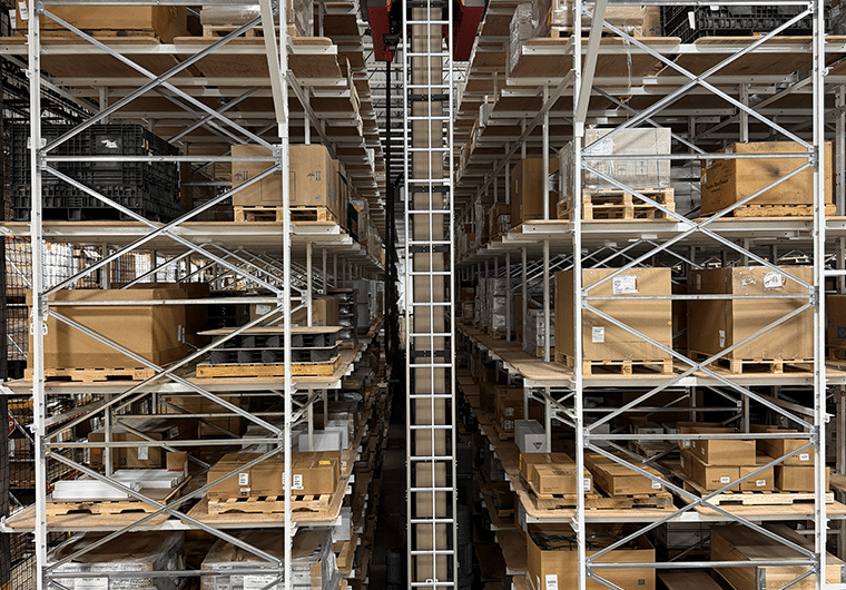 ASRS 2 Warehouse with tall metal racks loaded with pallets and boxes on both sides, seen from a central aisle with a ladder down the middle.