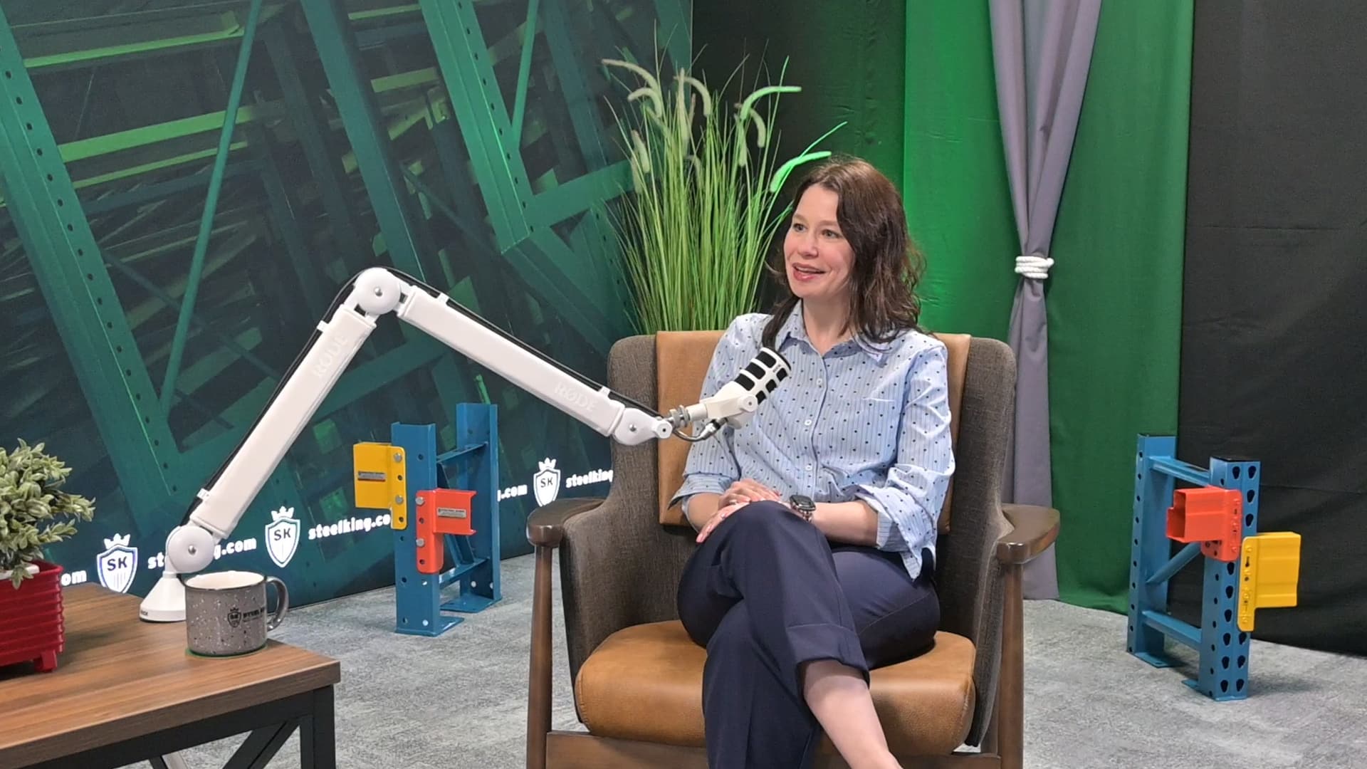 Woman in a blue polka-dotted shirt sits in a brown chair and speaks into a large white arm-mounted microphone in a studio setup with props and plants in the background.